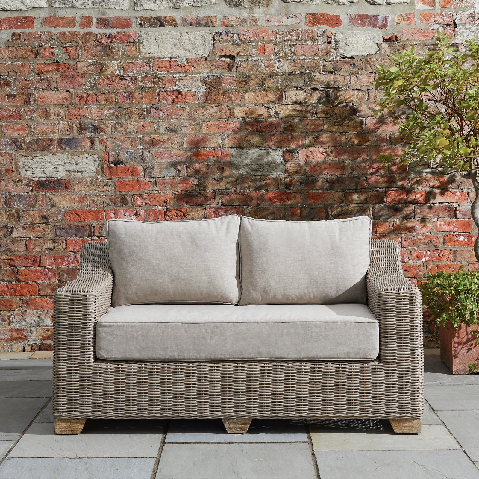 Wicker outdoor sofa with gray cushions against a brick wall.