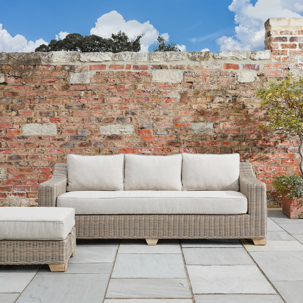 Outdoor wicker sofa with beige cushions against a brick wall.