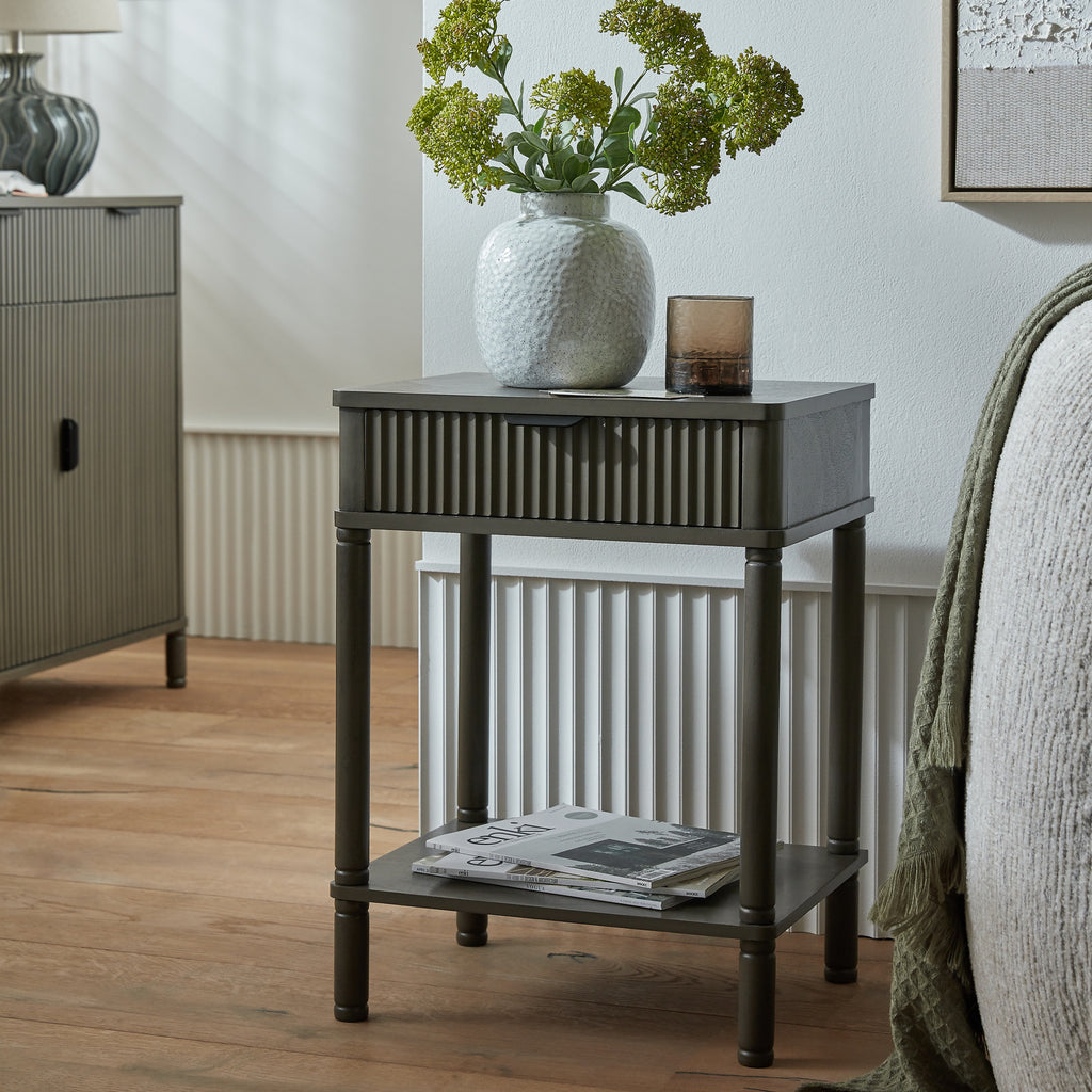 Small dark wooden side table with a white textured vase and greenery, next to a gray sofa and cabinet.
