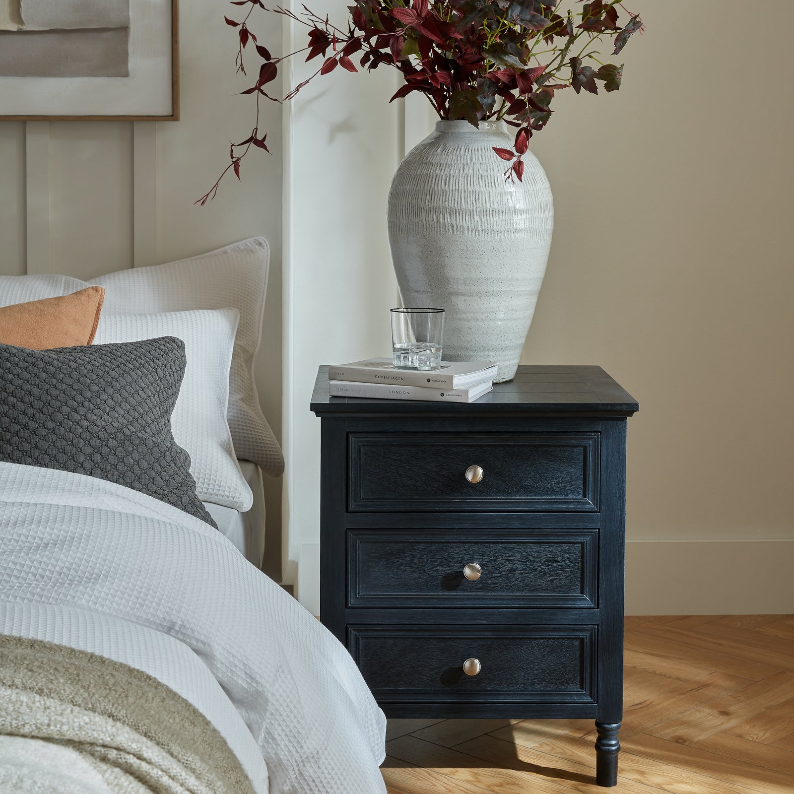 Nighstand with a vase and glass next to a bed with pillows